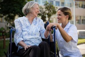 An elderly couple sits together in a cozy living room, each receiving personalized attention from a caregiver.